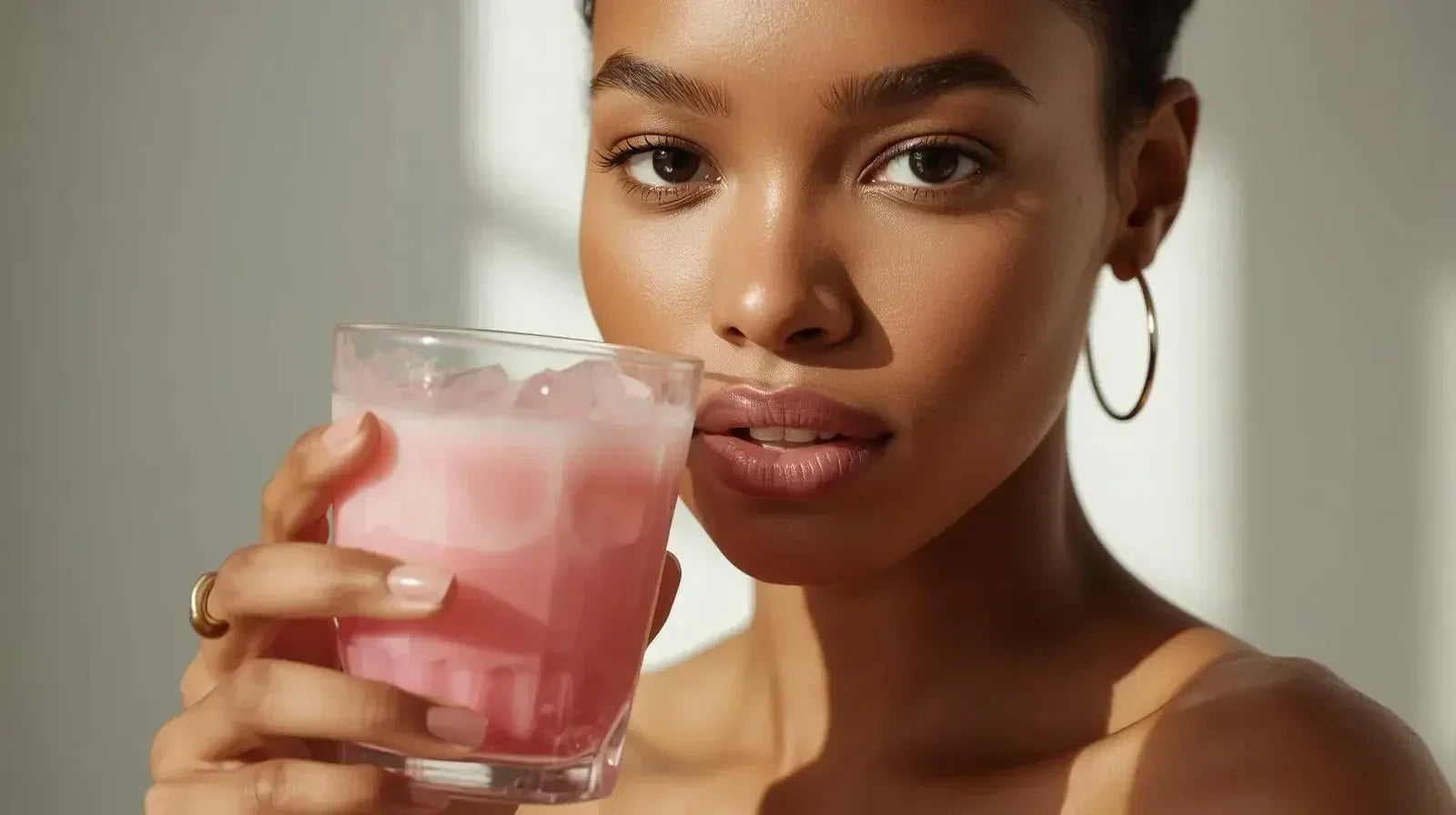 Close-up of woman with glowing skin holding a glass of pink collagen drink with ice
